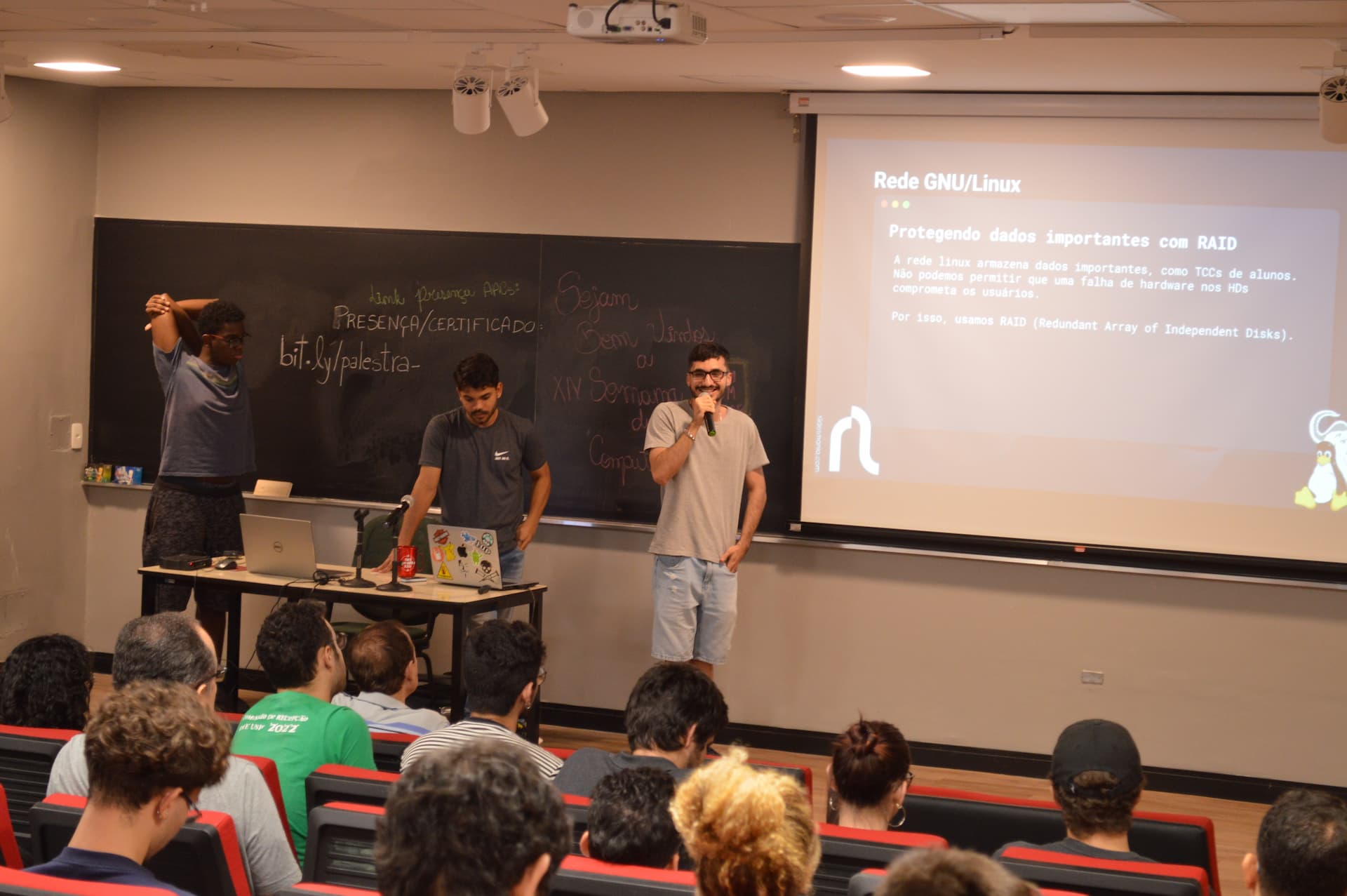 Três alunos apresentam o projeto da rede linux no projetor do auditório.